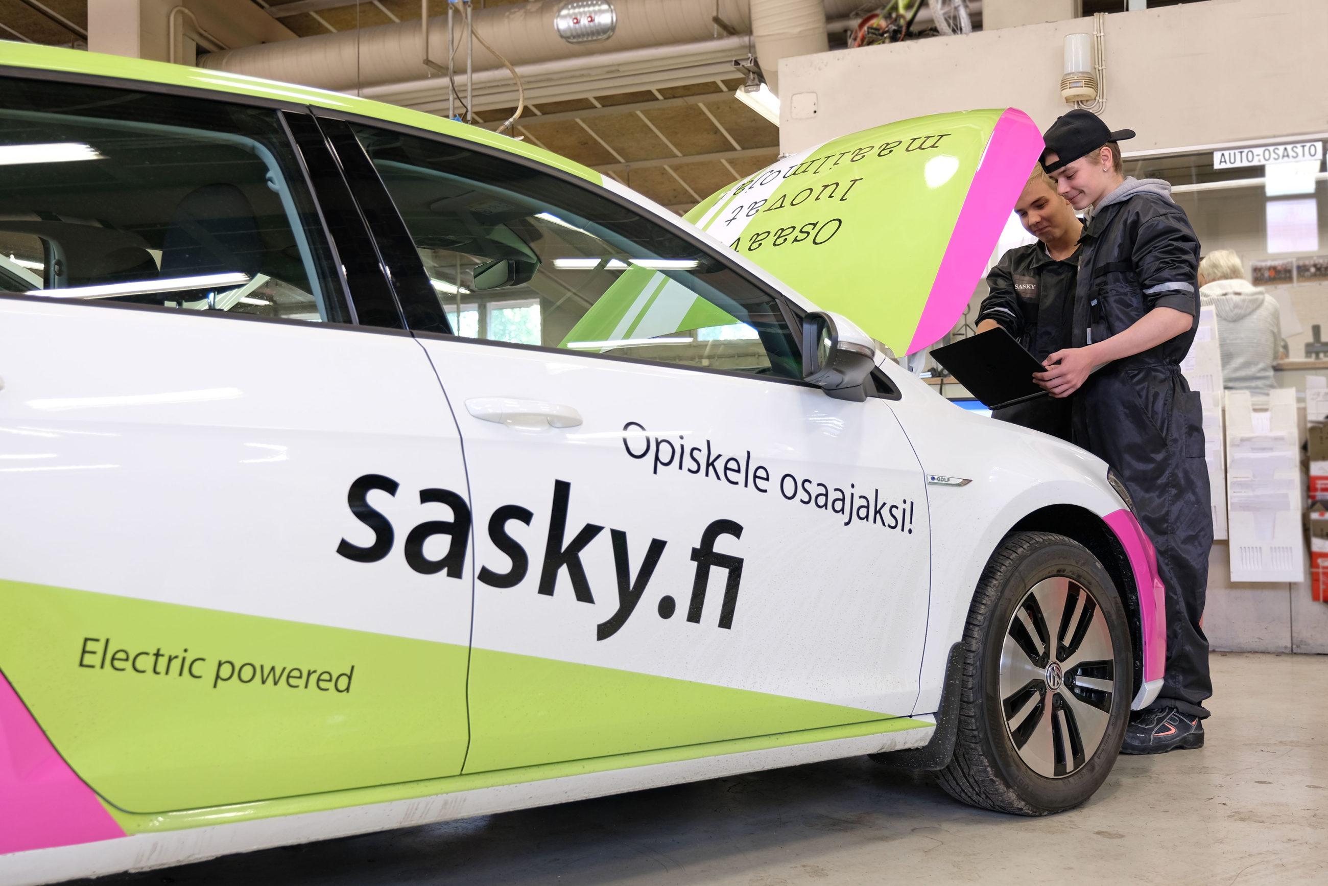 Kaksi henkilöä tutkii tietokone kädessään autoa, jonka konepelli on nostettu ylös, valko-vihreä-pinkin auton kyljessä on tekstit opiskele osaajaksi, sasky.fi ja electric powered.