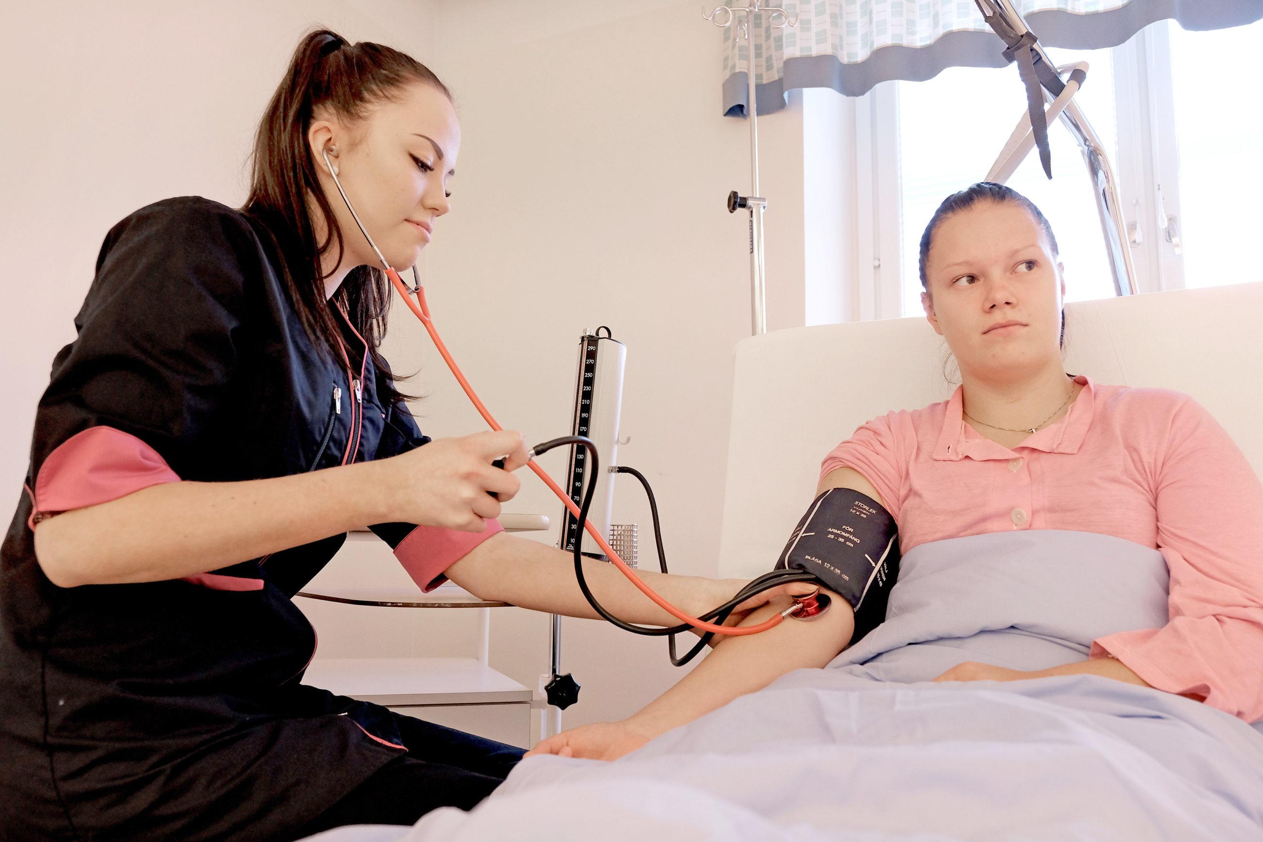 Henkilö mittaa verenpainetta toiselta henkilöltä, joka on sairaalasängyssä.