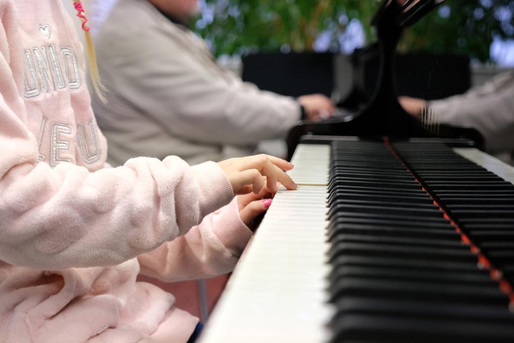 Etualalla lapsen kädet pianon koskettimilla, taustalla näkyy toisen henkilön kädet toisen pianon koskettimilla.