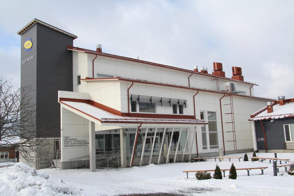 Valkoinen moderni koulurakennus, jossa vasemmalla reunalla harmaa tornimainen rakennelma, jonka seinässä sini-keltainen logo, Saskyn IDEA-kampus uudisrakennus Ikaalisissa.