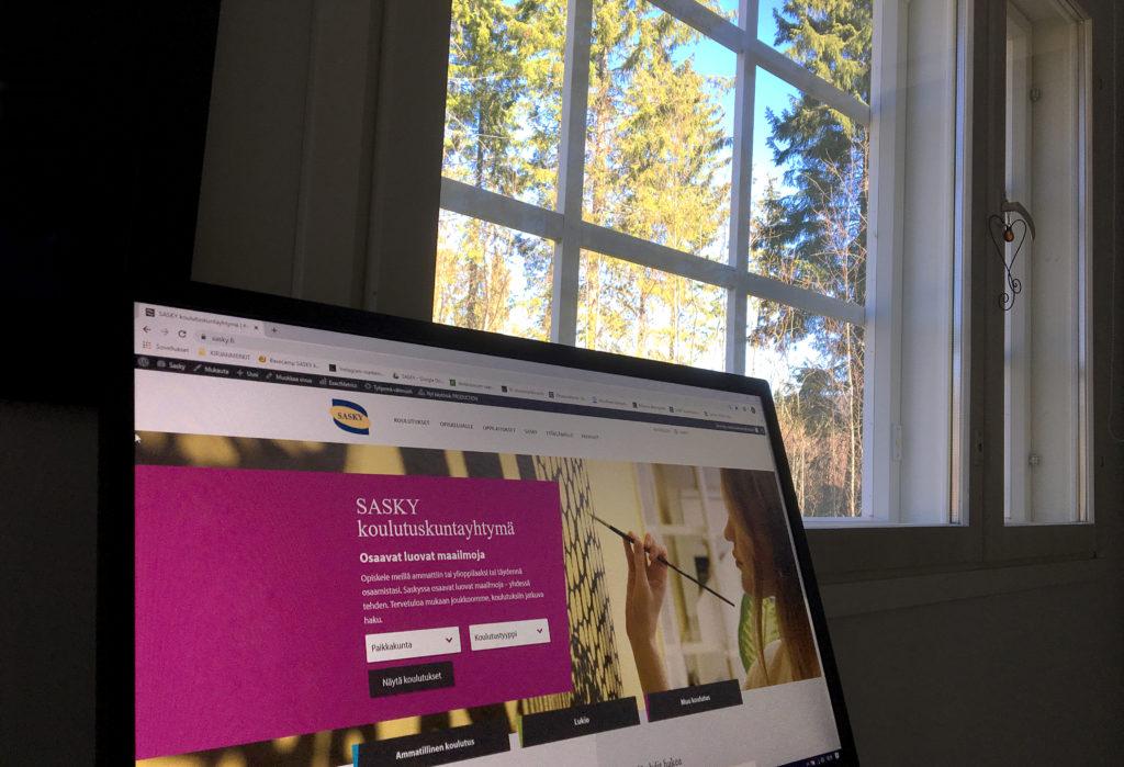 Tietokoneen ruudulla avoinna SASKY koulutuskuntayhtymän nettisivut, taustalla ikkuna, josta näkyy metsää ja sinistä taivasta.