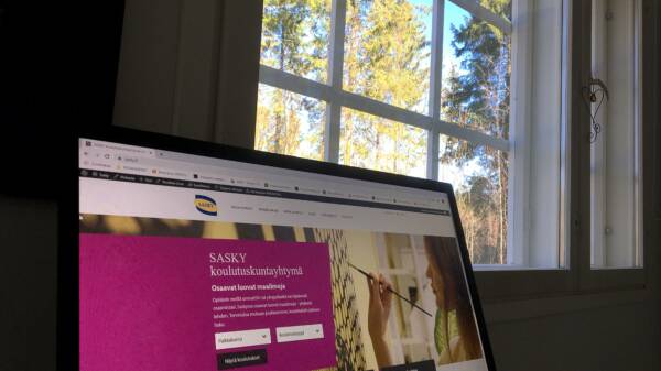 Tietokoneen ruudulla avoinna SASKY koulutuskuntayhtymän nettisivut, taustalla ikkuna, josta näkyy metsää ja sinistä taivasta.