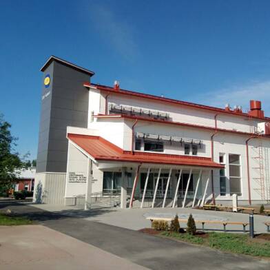 Valkoinen moderni koulurakennus, jossa vasemmalla reunalla harmaa tornimainen rakennelma, jonka seinässä sini-keltainen logo, Saskyn IDEA-kampus uudisrakennus Ikaalisissa, kesäinen kuva.
