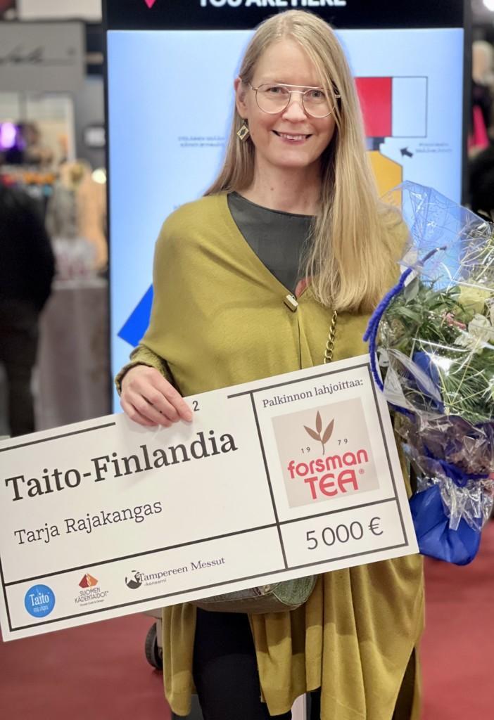 Hymyilevä, pitkähiuksinen nainen, jolla on kädessään kukkakimppu sekä kyltti, jossa teksti Taito-Finlandia Tarja Rajakangas.