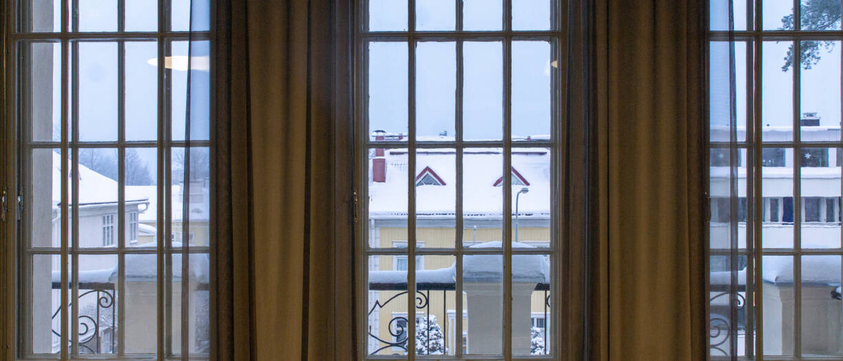 Näkymä ristikkoikkunasta ulos, osittain ikkunan edessä kellertävät verhot, ikkunan takana näkyy talvista kaupunkimaisemaa.