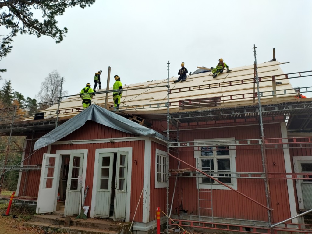 Punaisen puisen rakennuksen katolla viisi henkilöä työasuissa tekemässä kattoremonttia, rakennuksen ympärillä rakennustelineitä.