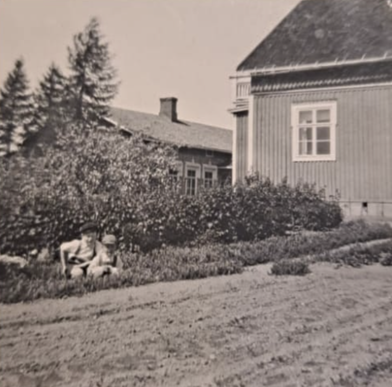 Mustavalkoinen vanha valokuva, jossa taustalla puisia rakennuksia ja etualalla kaksi lasta kasvimaan vieressä.