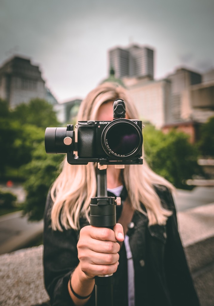 Vaaleahiuksinen henkilö pitää kädessään jalustalla olevaa kameraa niin, että kameran linssi on katsojaa päin ja henkilön kasvot jäävät kameran taakse piiloon, henkilön takana näkyy sumeana kaupunkimaisema.