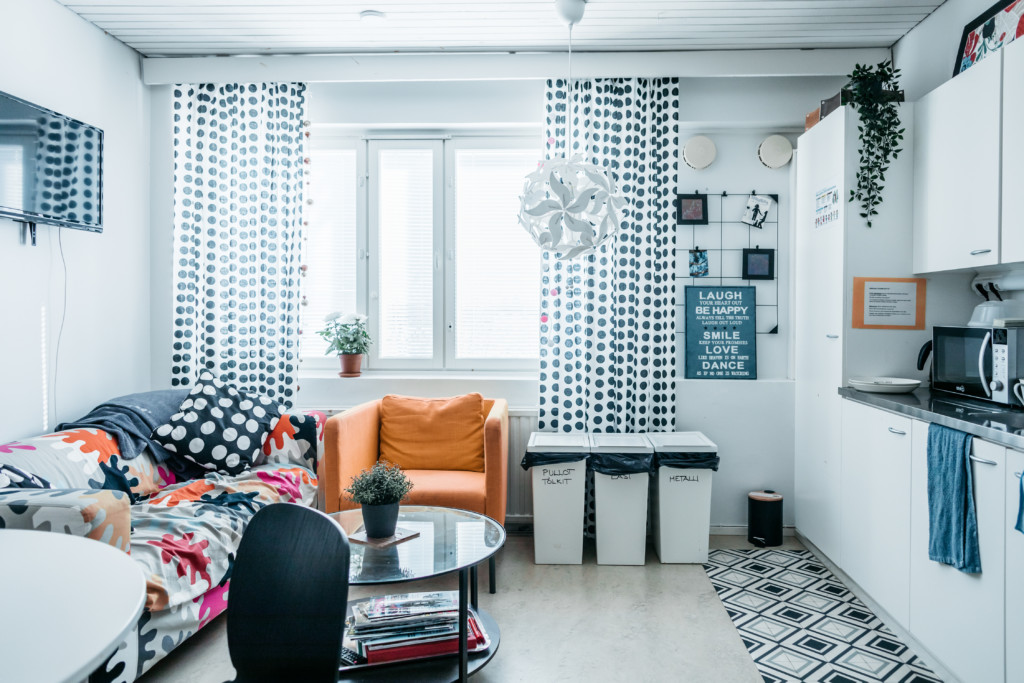 Kuva huoneesta, jossa värikkäällä kankaalla peitetty sohva, ikkunoissa mustavalkoiset verhot ja oikealla reunalla minikeittiö.