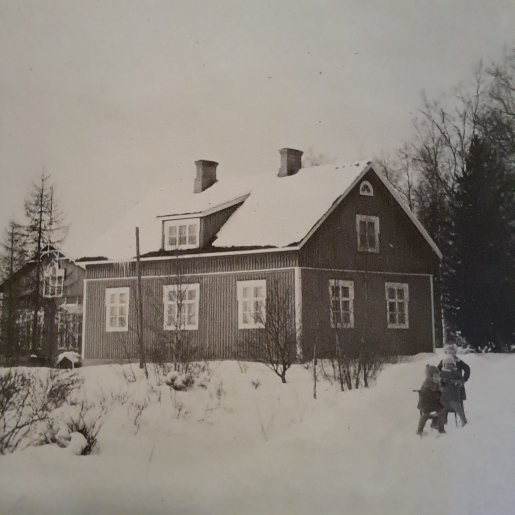 Mustavalkoinen vanha valokuva puutalosta talvimaisemassa.