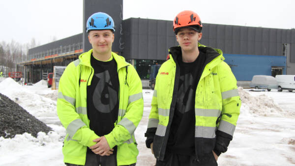 Kaksi keltaisiin työtakkeihin pukeutunutta henkilöä rakennustyömaan edessä pihalla, toisella sininen työmaakypärä ja toisella punainen.
