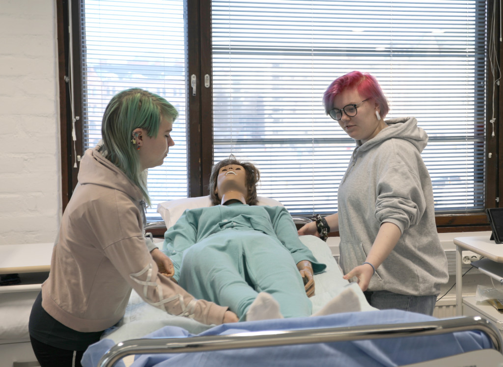 Kaksi henkilöä harjoittelee lähihoitajan työtä hoitotyön harjoittelussa käytettävän nuken kanssa sairaalavuoteen ääressä.