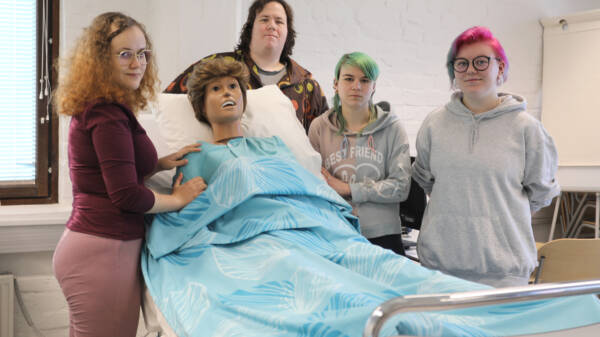 Neljä henkilöä sairaalavuoteen ympärillä, vuoteessa turkoosin lakanan alla hoitotyön harjoittelussa käytettävä nukke.