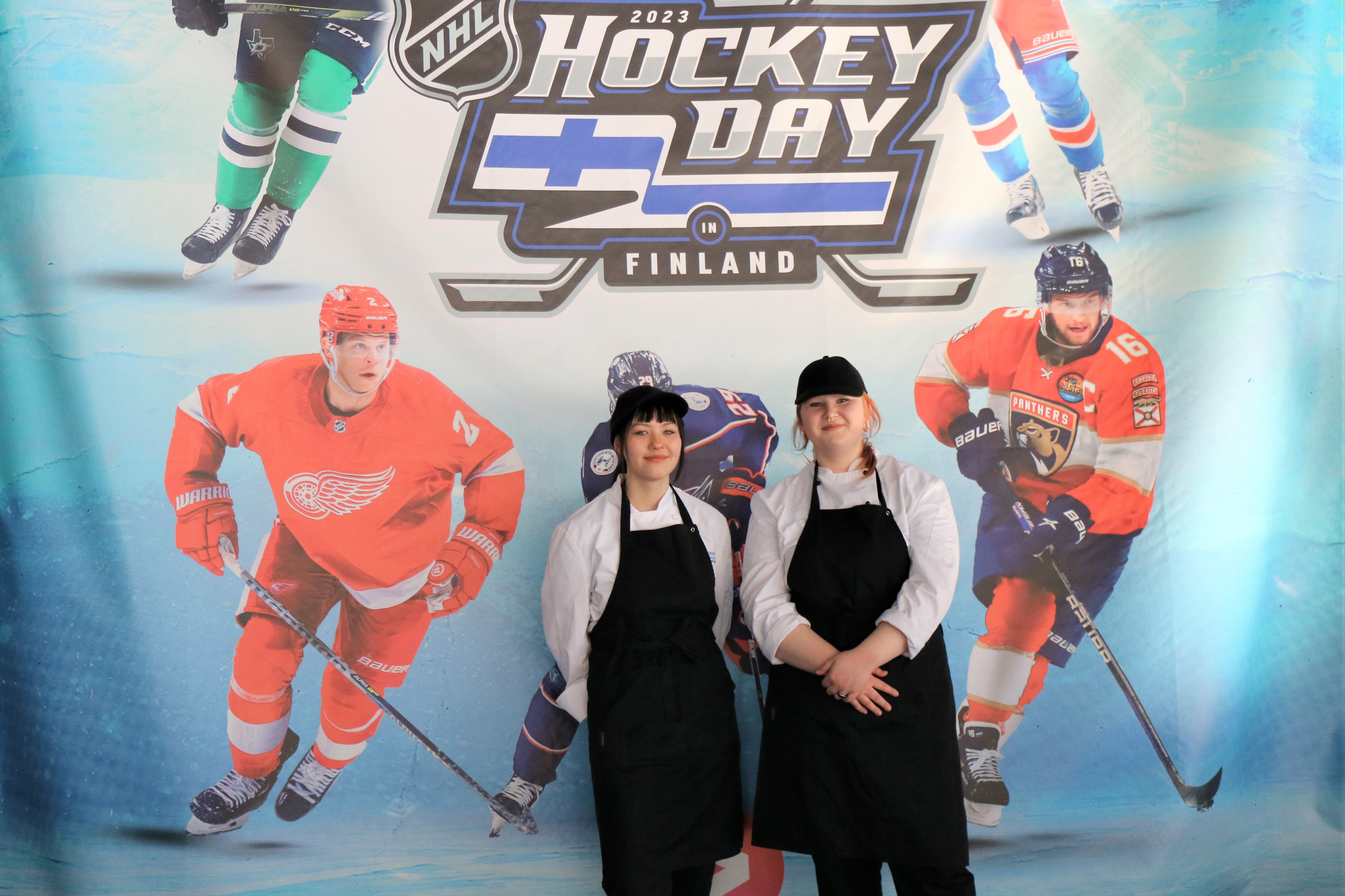 Kuvassa Hockey Day -tausta jossa tapahtuman otsikko sekä kuvia jääkiekkoilijoista. Etualalla Milo ja Tuulia yllään mustat essut.