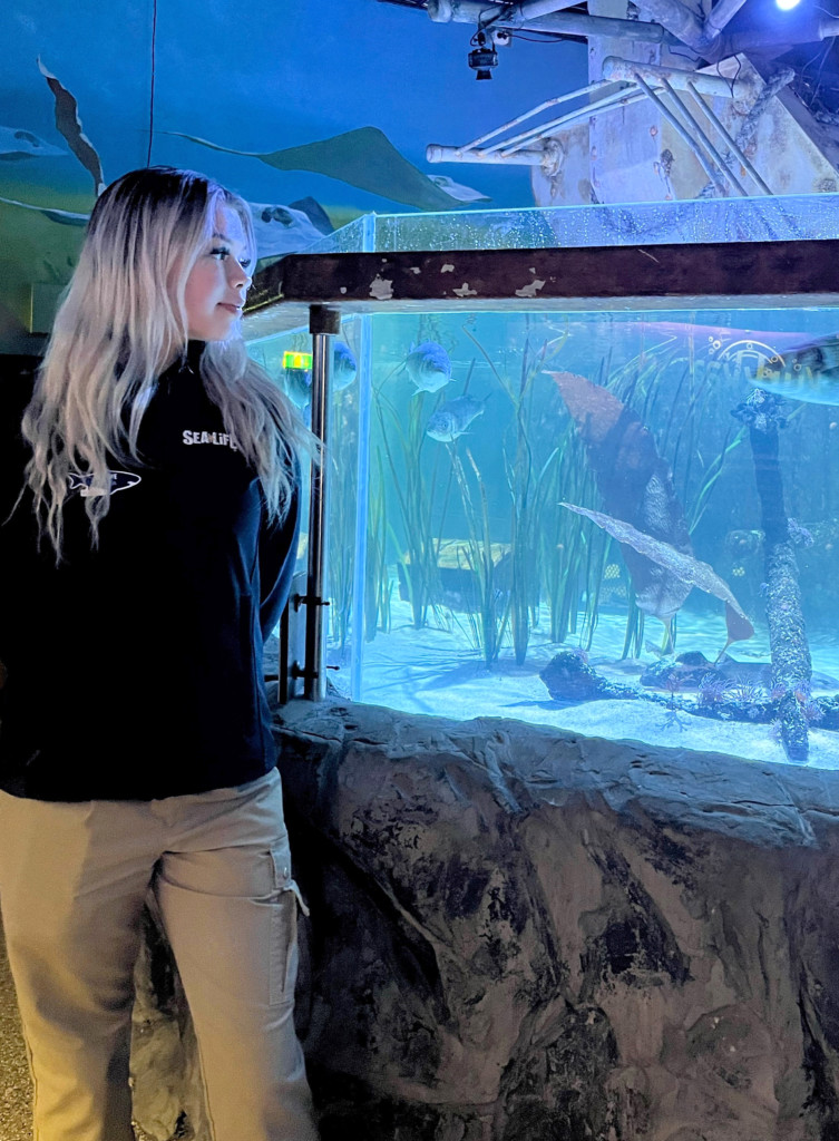 Vaaleahiuksinen henkilö tummassa hupparissa ja vaaleissa housuissa seisoo ison akvaarion edessä.