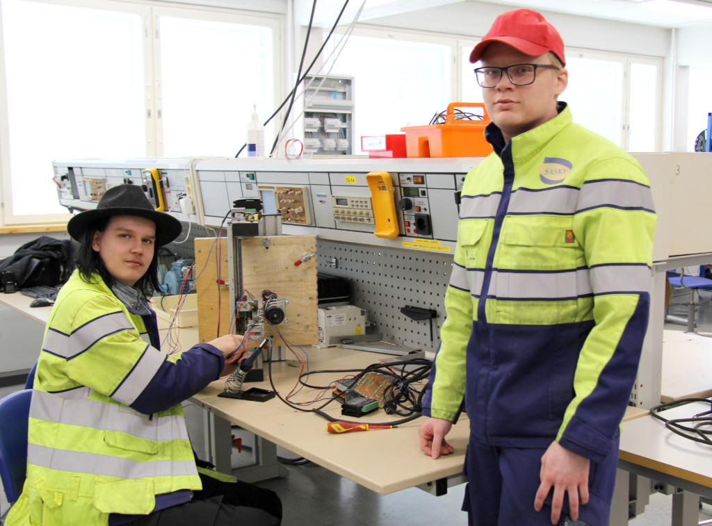 Kaksi henkilöä työpöydän äärellä korjaamassa sähkölaitetta, molemmilla keltaiset työtakit, toisella päässään musta hattu ja toisella punainen lippalakki.