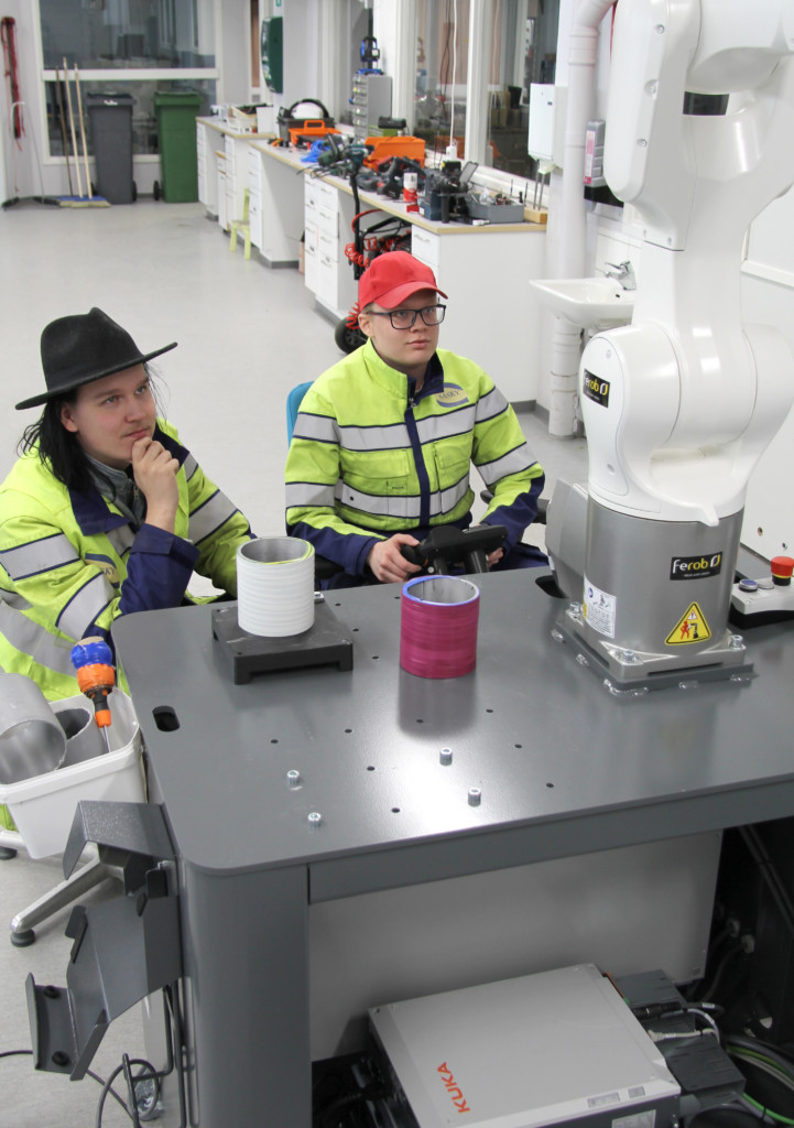 Kaksi henkilöä työpöydän äärellä ohjaamassa robotin toimintaa, molemmilla keltaiset työtakit, toisella päässään musta hattu ja toisella punainen lippalakki.