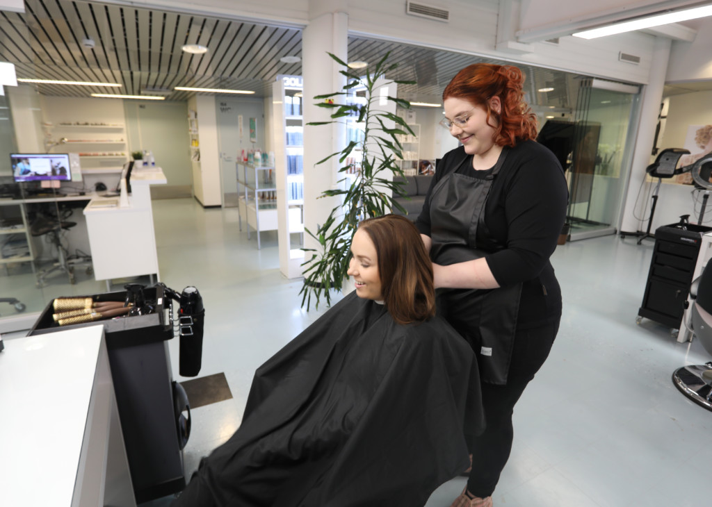Punertavahiuksinen, silmälasipäinen kampaaja, jolla on musta paita ja musta esiliina, laittaa kampaamotuolissa istuvan, pitkähiuksisen asiakkaan hiuksia, taustalla näkyy kampaamomiljöötä.
