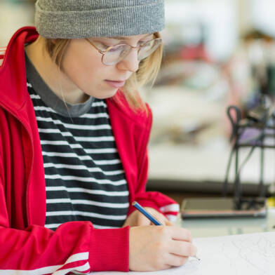 Silmälasipäinen opiskelija, jolla harmaa pipo ja punavalkoinen verkkaritakki piirtää sisustuslasin suunnitelmaa paperille.