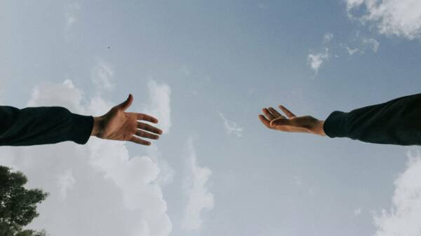 Kaksi kättä kurottelevat toisiaan, taustalla näkyy sininen taivas ja pilviä.