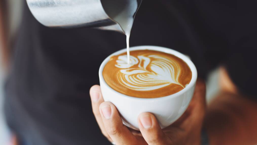 Henkilöllä kädessä kahvikuppi, johon hän valuttaa vaahdotettua maitioa ja kahvin pintaan muodostuu lehtikuvio