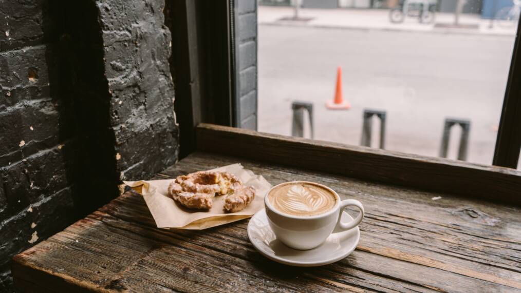 Kuppi kahvia sekä leivonnainen kahvilan pöydällä ikkunan edessä.