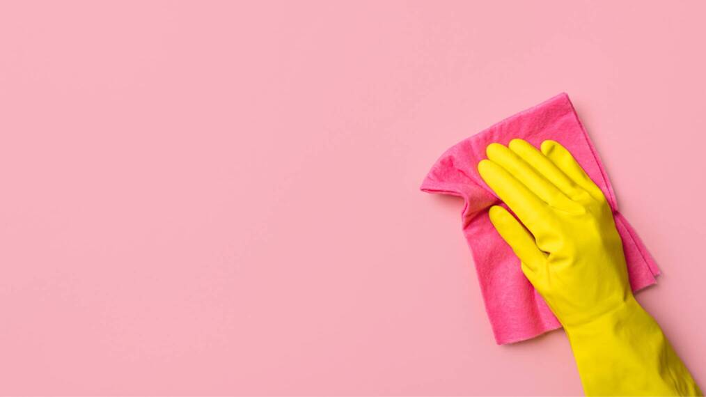 Vaaleanpunaisella taustalla vaaleanpunainen siivousliina, jota pitelee keltaiseen kumihansikkaaseen puettu käsi.