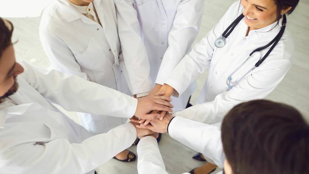 Lääkärit seisovat kehässä ja pitävät käsiä päällekkäin kuvan keskellä.