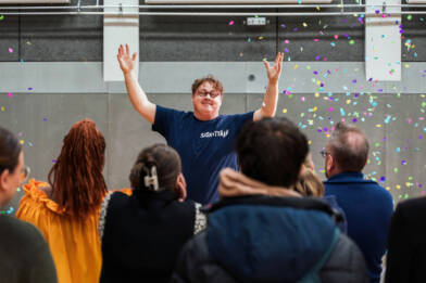 Tummansiniseen Saskyttäjä-t-paitaan pukeutunut henkilö kädet ilmassa ympärillään värikkäitä konfetteja, etualalla yleisöä selin kameraan.