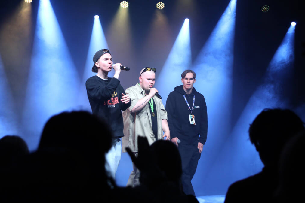 Kolme henkilöä lavalla kohdevaloissa, kaksi vasemmanpuoleista laulaa mikrofoniin, etualalla näkyy yleisön tumma silhuetti.