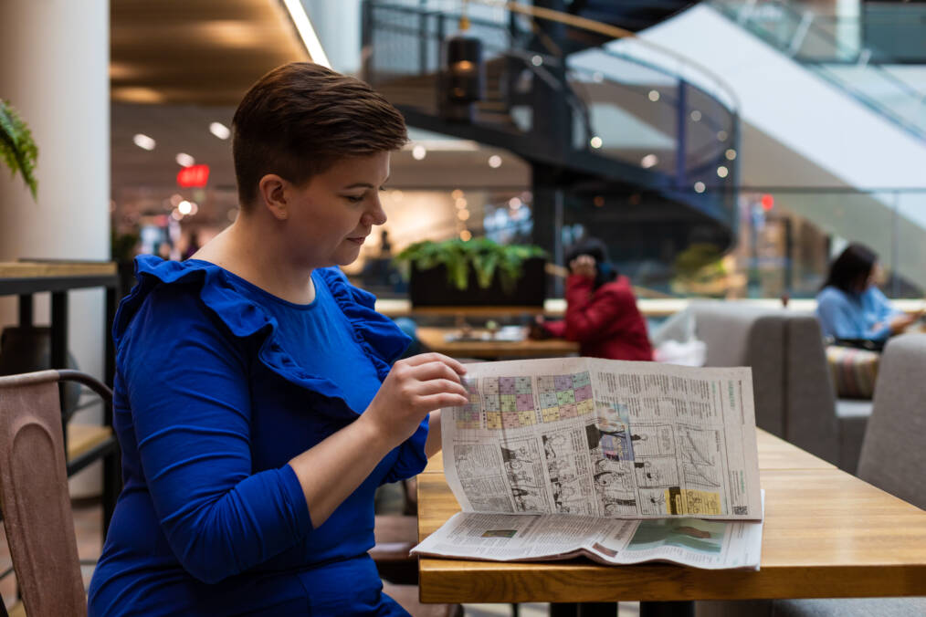 Henkilö istuu kahvilassa ja lukee sanomalehteä. Henkilöllä on yllään kirkkaan sininen mekko. Taustalla liiketiloja ja kierreportaat.