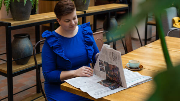 Henkilö istuu kahvilassa ja lukee sanomalehteä. Henkilöllä on yllään kirkkaan sininen mekko. Tilassa paljon viherkasveja.
