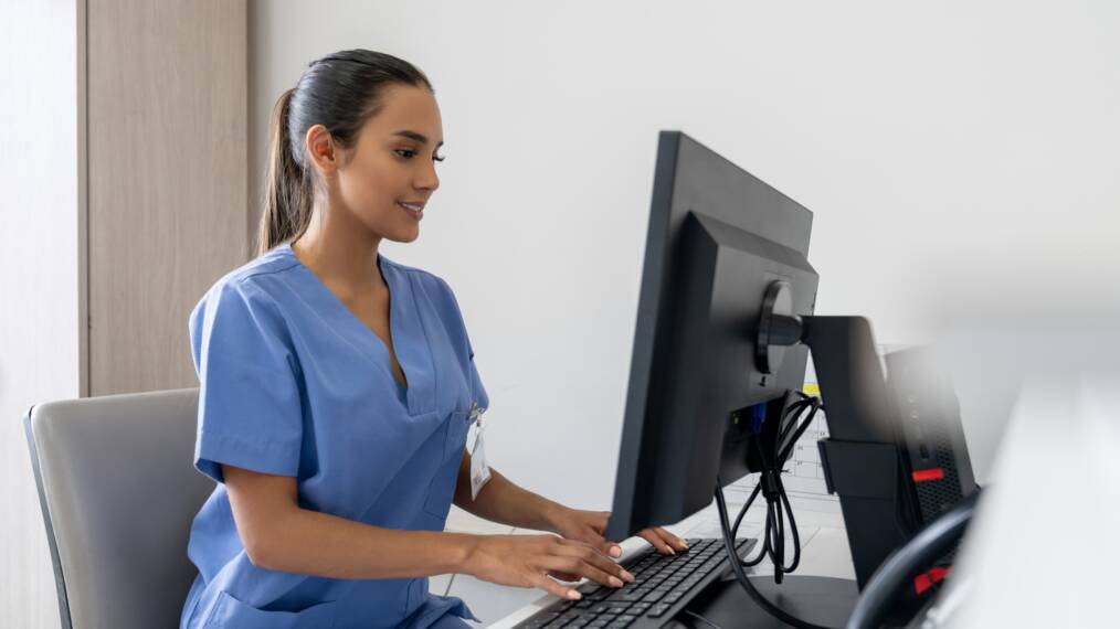 Kuvassa tummahiuksinen henkilö pukeutuneena sairaalan siniseen työasuun. Henkilö istuu työpöydän ääressä ja käyttää tietokonetta.