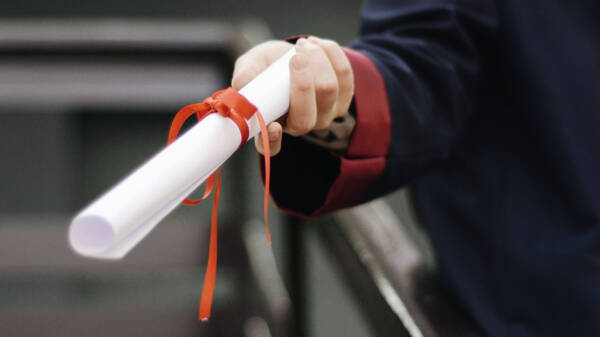 Lähikuva henkilön kädestä, jossa on valkoinen, punaisella silkkinauhalla sidottu paperirulla.