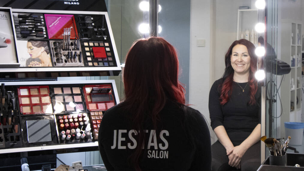 Hymyilevä punahiuksinen henkilö mustassa paidassa katsoo peiliin. Taustalla on telineessä erilaisia meikkituotteita.
