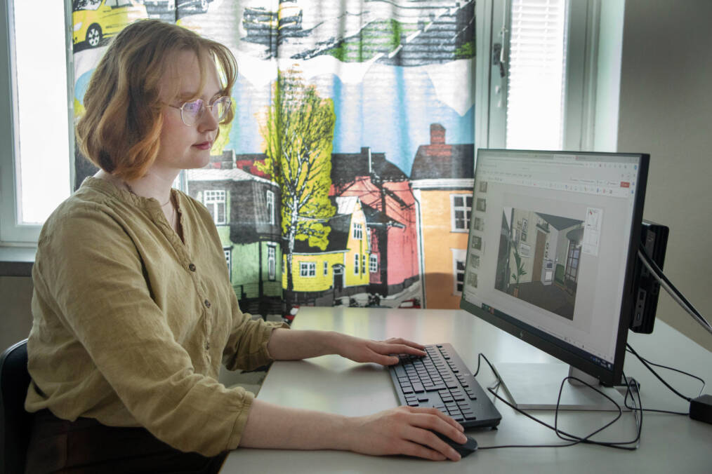 Henkilö istuu tietokoneen ääressä ja työskentelee huonesuunnitelman parissa, hänellä on vaaleat hiukset ja silmälasit ja yllään vaaleankelletävä paita, taustalla on värikäs verho, jossa on talojen kuvia.