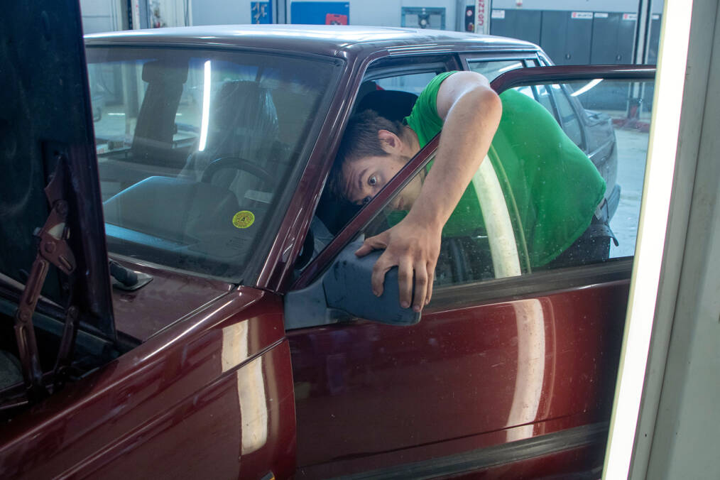 Henkilö vihreässä t-paidassa säätää auton sivupeiliä autokorjaamossa, hän on kumartunut auton sisäpuolelle, pitäen kiinni peilistä ulkopuolelta, taustalla näkyy työpajan sisätilat ja auton konepelti on auki.