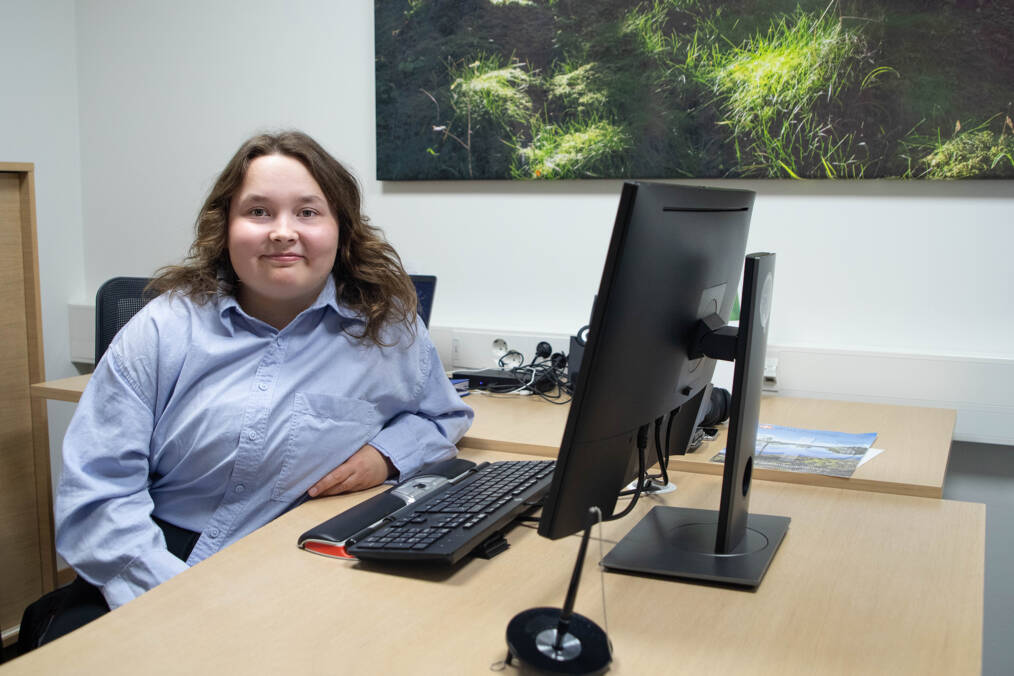 Vaaleansiniseen paitaan pukeutunut, tumma- ja kiharahiuksinen henkilö istuu toimistossa työpöydän ääressä tietokoneen vieressä, taustalla näkyy seinälle ripustettu maisemakuva, henkilö katsoo suoraan kohti kameraa ja hymyilee.