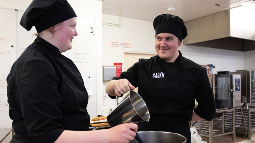 Kaksi mustiin kokintakkeihin pukeutunutta nuorta keittiöympäristössä kattilan äärellä