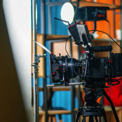Ammattimainen videokamera jalustalla studiossa tai tuotantotilassa, taustalla näkyy hyllykköjä ja epäsuoraa valaistusta.