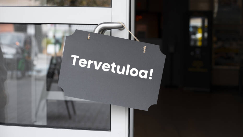 Avoinna olevassa ovessa roikkuu harmaa kyltti, jossa on teksti tervetuloa.