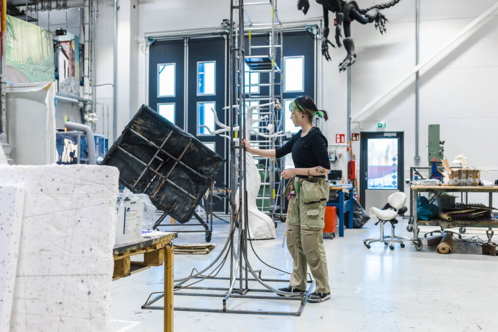 Henkilö seisoo työpajassa metallisen veistoksen vieressä. Hän katselee rakenteen yksityiskohtia, ja taustalla näkyy erilaisia työvälineitä ja suuria taideteoksia.