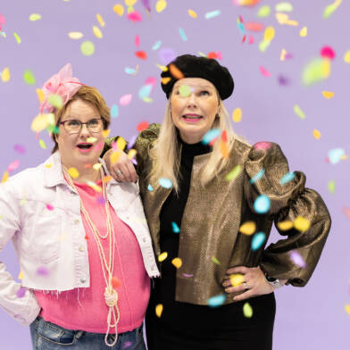 Kaksi iloista opinto-ohjaajaa poseeraa värikkään konfettisateen keskellä, toinen pukeutuneena pinkkiin ja toinen kultaisiin sävyihin.