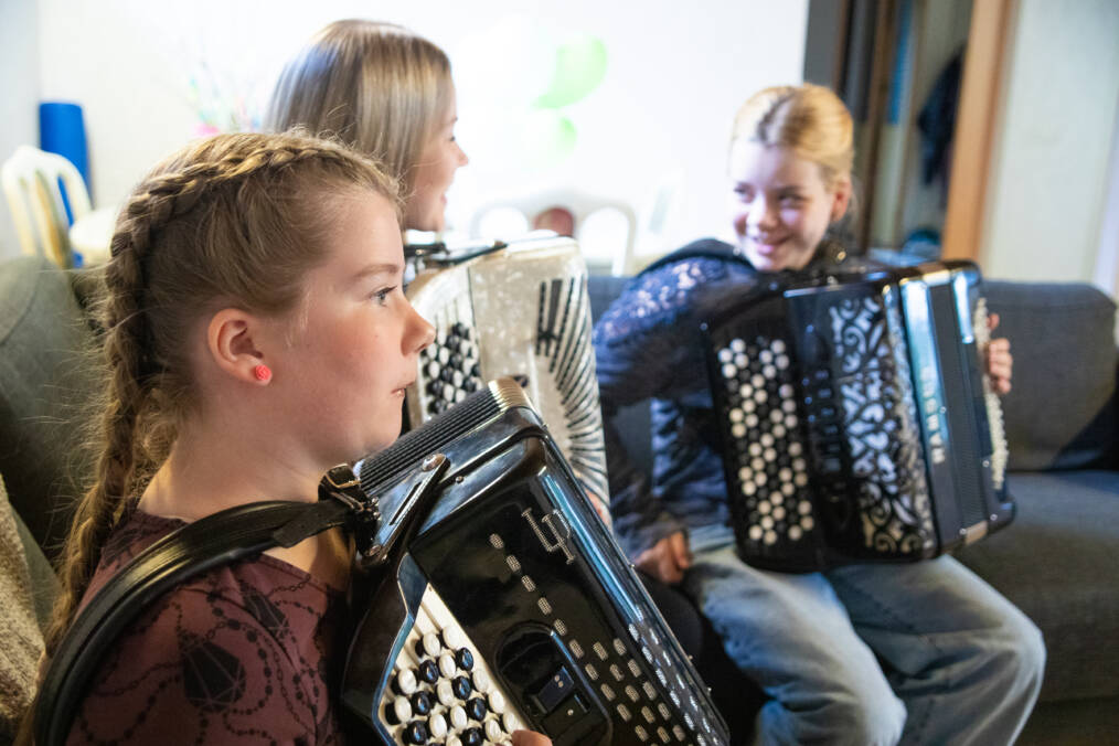 Etualalla lettipäinen tyttö soittaa haitaria, taustalla näkyy samalla sohvalla istumassa kaksi muutakin haitarinsoittajaa.