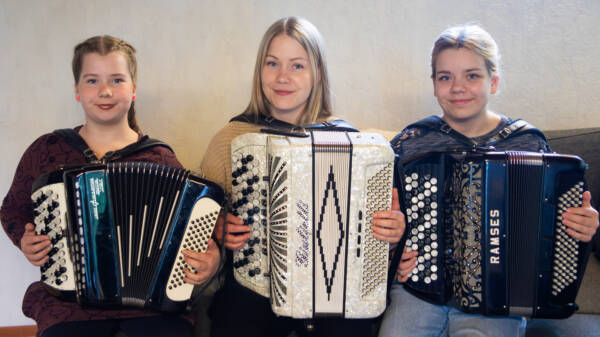 Kolme henkilöä istuu vierekkäin sohvalla ja soittaa haitaria katsoen samalla hymyillen kameraan.