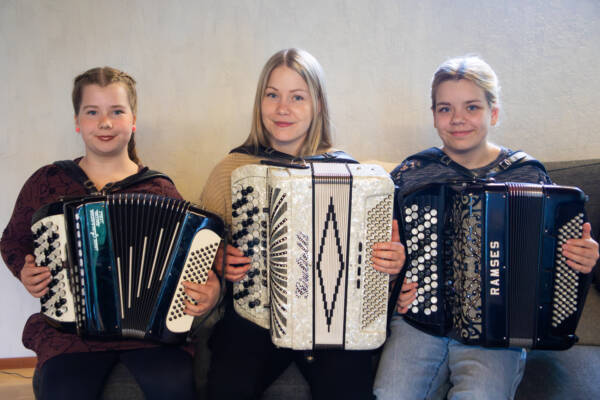 Kolme henkilöä istuu vierekkäin sohvalla ja soittaa haitaria katsoen samalla hymyillen kameraan.