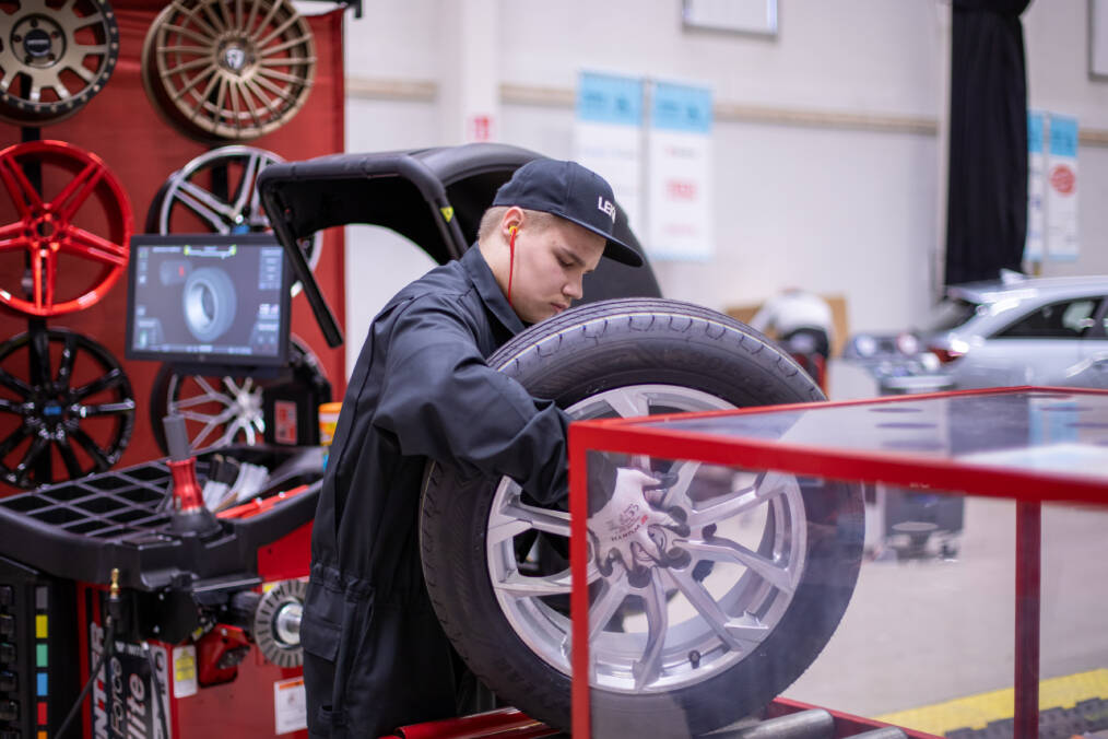 Tummansiniseen haalariin ja lippalakkiin pukeutunut henkilö nostaa isoa autonrengasta tason päälle, takana näkyy kilpailualue sekä autonvanteita punaista taustaa varten.