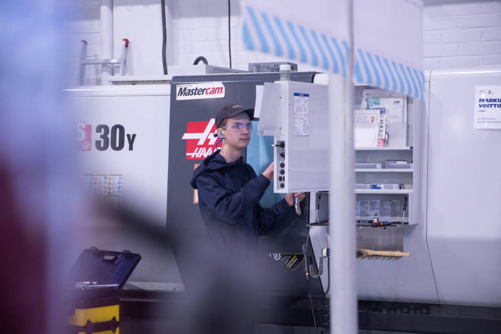 Tummansiniseen huppariin ja lippalakkiin pukeutunut henkilö tekee töitä CNC-työstökeskuksella.