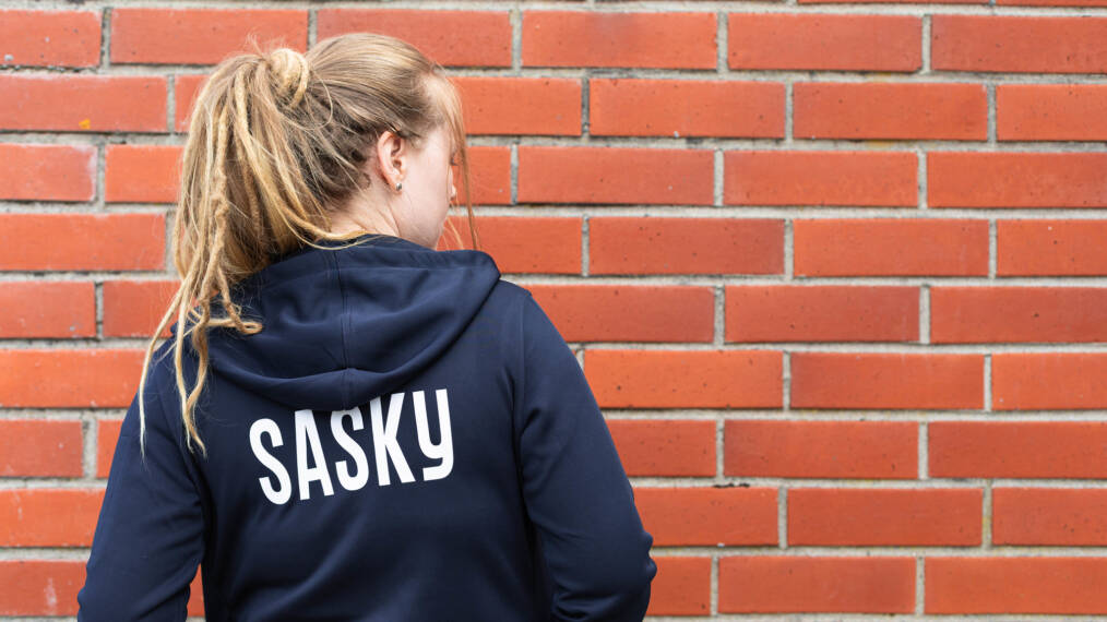 Rastatukkainen henkilö seisoo tiiliseinän edessä selin kameraan, hänellä on päällään tummansininen huppari, jonka selässä on teksti Sasky.