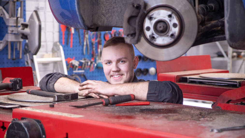 Autonasentaja työssään korjaamolla auton nosturin alla.